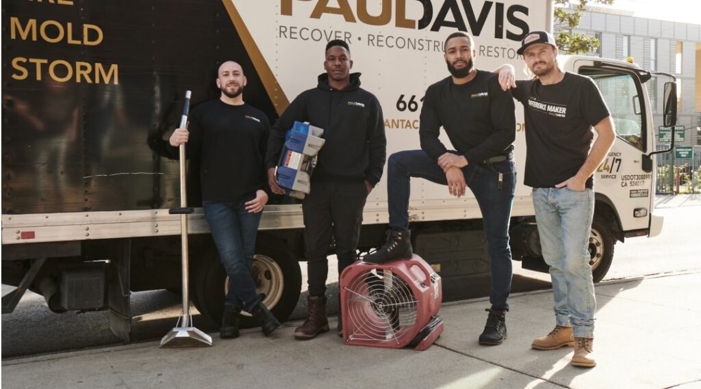 Four technicians from Paul Davis Restoration of Portland / Vancouver stand together beside a branded service truck, posing with restoration equipment including a floor scraper and an air mover fan on a city sidewalk.